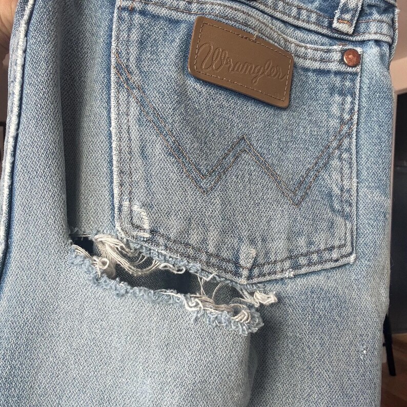May include: Close-up of light blue denim jeans with a distressed, ripped design on the back pocket area. The jeans feature a brown leather Wrangler patch and a classic pocket design.