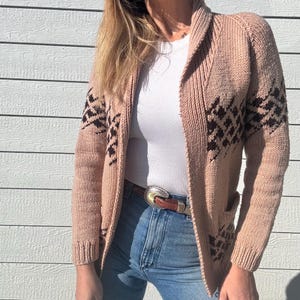 May include: A pink knitted cardigan with a brown geometric pattern. The cardigan has a shawl collar and long sleeves. The model is wearing the cardigan with a white t-shirt and blue jeans.