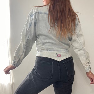 May include: A light blue denim jacket with a floral embroidery detail on the back. The jacket is cropped and has a slightly fitted silhouette.