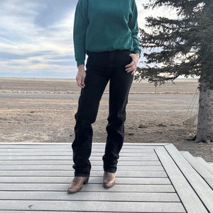 May include: A person wearing a dark green sweatshirt and black jeans. The person is also wearing brown cowboy boots. The outfit is casual and suitable for outdoor activities. The background includes a wooden deck, a field, and a cloudy sky.