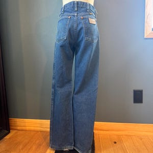 May include: Blue denim jeans with a classic five-pocket design. The jeans have a straight leg fit and a brown leather patch on the back pocket. The jeans are displayed on a mannequin.