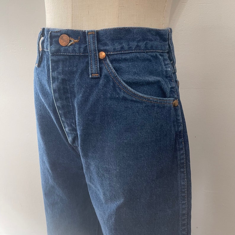 Pu&ograve; includere: Un paio di jeans in denim blu scuro con un bottone e rivetti in rame. I jeans hanno uno stile classico a cinque tasche.