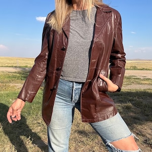 May include: A brown leather jacket with a button-up front and a collar. The jacket has a relaxed fit and is worn over a grey t-shirt and blue jeans.