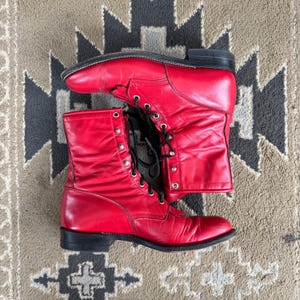May include: A pair of bright red leather lace-up boots featuring black laces, silver-toned metal eyelets, and a black stacked heel. The boots have a smooth finish with visible creasing on the vamp and shaft, and a rounded toe design.