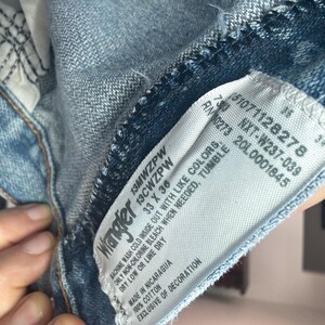 May include: Close-up of a light blue denim jeans tag. The tag reads "Wrangler" and includes size information: 33 x 36. Additional text includes washing instructions and material composition. The jeans are made in Nicaragua.