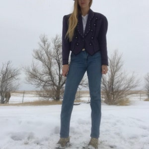 May include: A purple cropped jacket with a pointed collar, silver buttons, and a decorative design on the front. The jacket is worn over a white shirt and blue jeans.