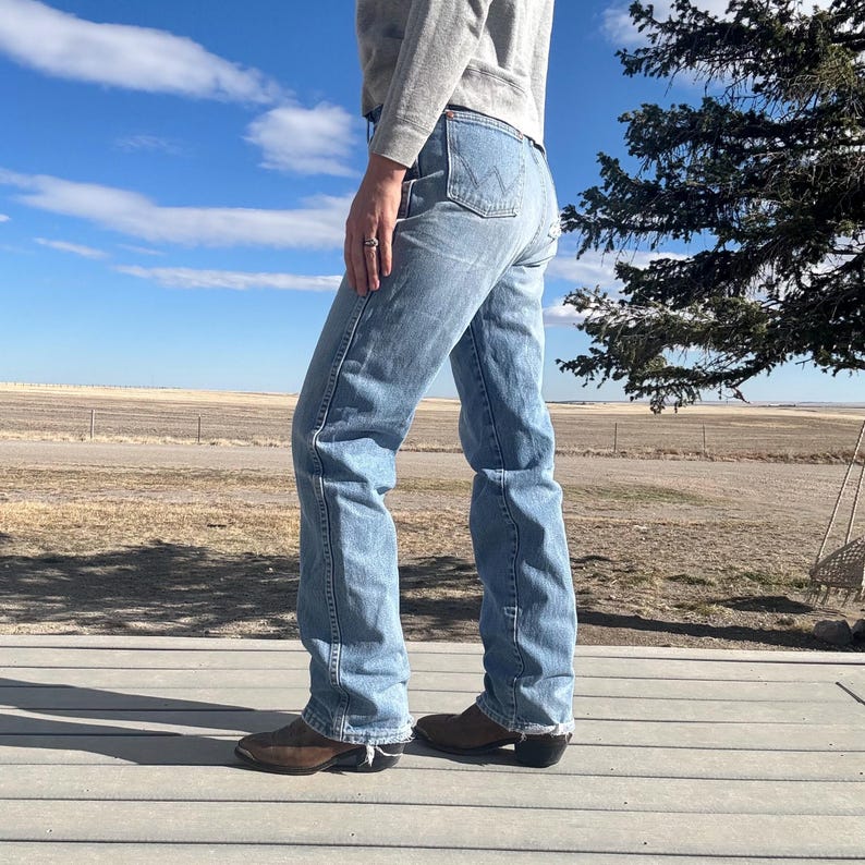 May include: Light blue denim jeans with a classic fit, featuring a high waist and a straight leg. The jeans are paired with brown cowboy boots. The background shows a blue sky with clouds and a rural landscape.