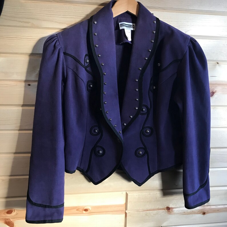 May include: A purple cropped jacket with black trim and silver studs. The jacket has a pointed collar and a decorative design on the front.