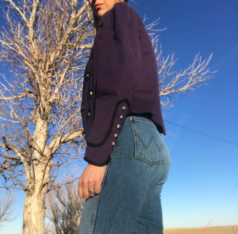 May include: A purple jacket with black trim and silver buttons. The jacket has a fitted silhouette and is cropped at the waist. The person wearing the jacket is also wearing blue jeans.