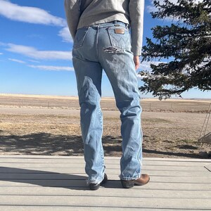 May include: Light blue denim jeans with distressed details, featuring a classic five-pocket design and a brown leather patch on the back. The jeans have a straight leg fit and are worn with brown leather boots.