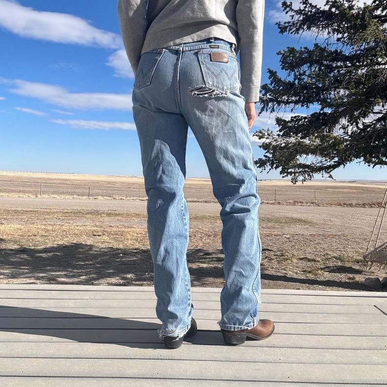 May include: Light blue denim jeans with distressed details, featuring a classic five-pocket design and a brown leather patch on the back. The jeans have a straight leg fit and are worn with brown leather boots.