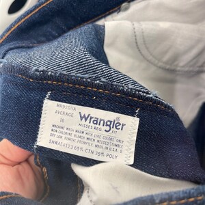 May include: A white Wrangler jeans care label with blue text. The label says "Misses Reg. Fit" and includes washing instructions. The label also says "MR920NA Average 18 5HWAE4123 65% CTN 35% POLY".
