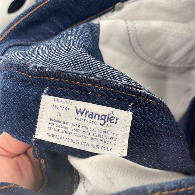 May include: A white Wrangler jeans care label with blue text. The label says "Misses Reg. Fit" and includes washing instructions. The label also says "MR920NA Average 18 5HWAE4123 65% CTN 35% POLY".