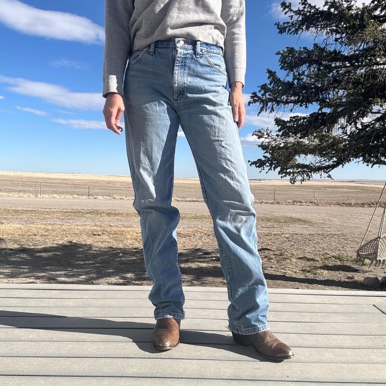 May include: Light blue denim jeans with a straight leg fit. The jeans have a classic five-pocket design and are worn with brown leather boots. The person is wearing a gray long-sleeve shirt. The background shows a rural landscape under a blue sky.