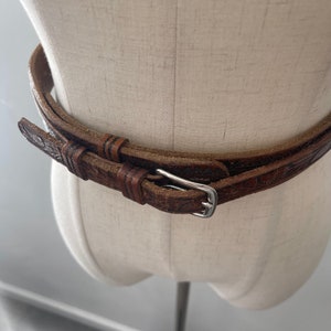 May include: A brown leather belt with an intricate embossed pattern and a silver buckle. The belt is double-layered and appears to be handmade.