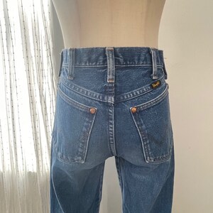 以下が含まれることがあります： ハイウエストでクラシックな5ポケットスタイルのブルーデニムジーンズ。ジーンズのバックポケットには銅製の鋲が付き、右側のバックポケットには「Wrangler」と書かれた小さな革のパッチが付いています。