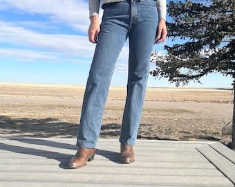 May include: A person wearing a gray sweatshirt and blue jeans with brown leather boots. The jeans are a light blue wash and have a straight leg. The boots have a pointed toe and a small heel. The background is a field and a blue sky.