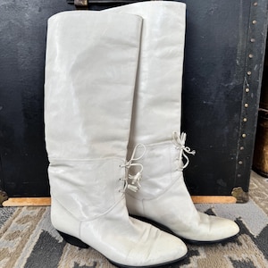 Puede incluir: Un par de botas altas hasta la rodilla de color blanco roto con acabado liso, punta redondeada y tacón bajo negro. Las botas incluyen un detalle de cordones decorativo con pequeñas borlas situadas en la parte frontal del tobillo.