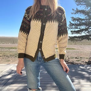 Könnte beinhalten: Cremefarbene Strickjacke mit Knöpfen und olivgrünen und roten geometrischen Mustern auf den Schultern und der oberen Brust. Der Pullover hat ein gepunktetes Muster auf der Vorderseite und den Ärmeln, mit olivgrüner Einfassung und silbernen Knöpfen. Die Person trägt eine blaue Jeans.