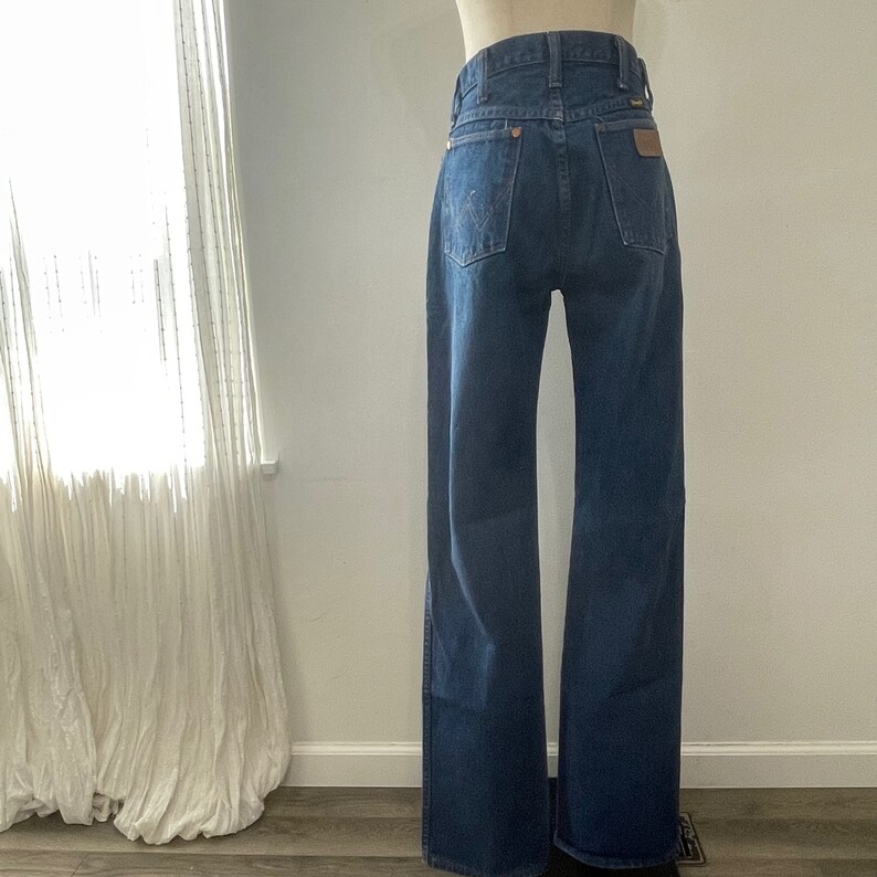 Pu&ograve; includere: Un paio di jeans in denim blu scuro con gamba larga. I jeans hanno vita alta e un design classico a cinque tasche. Le tasche posteriori hanno un toppa in pelle marrone con cuciture.