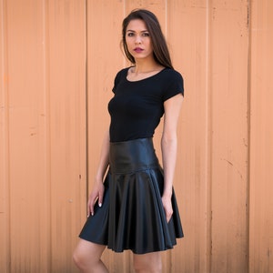 May include: A woman wearing a black short-sleeved shirt and a black leather skirt. She is standing in front of a peach-colored wall.