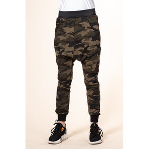 May include: Camouflage print pants with a black waistband and black trim at the ankles. The pants have a loose fit and a unique design with a large pocket on the right leg.