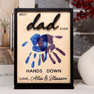 May include: A wooden plaque with a black frame and stand. The plaque reads "BEST dad EVER" in black raised letters. Below are blue and purple handprints and the words "HANDS DOWN LOVE, Atlas & Blossom."