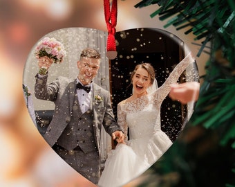 May include: A heart-shaped ornament with a photo of a bride and groom exiting a building, confetti falling around them. The ornament is hanging from a red ribbon on a green Christmas tree.