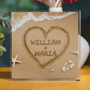 May include: A clear acrylic frame with a heart drawn in the sand, with the names "William + Maria" written inside the heart. The frame also contains a starfish, seashells, and a wave of sand.