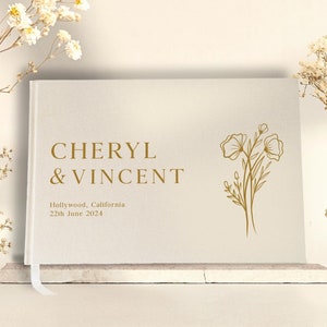 Puede incluir: Un libro de visitas de boda color crema con letras doradas que dice "CHERYL & VINCENT". El libro incluye la fecha y el lugar de la boda. Un dise&ntilde;o floral dorado est&aacute; en el lado derecho. Una cinta blanca de marcador es visible.