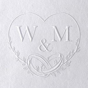 May include: A white embossed design on textured paper. The design features a heart shape with the letters "W" and "M" separated by an ampersand. Below the letters are two intertwined rings, surrounded by a leafy wreath.