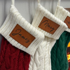 May include: Three white knitted Christmas stockings with brown leather name tags. The stockings are hung on a white fireplace mantel. The names on the tags are "James", "Mary", and "Thomas".