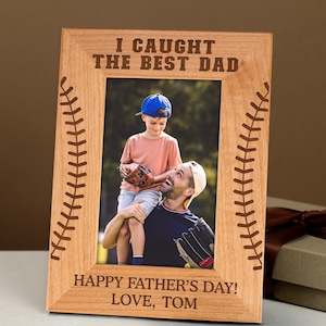 May include: A wooden picture frame with the text "I CAUGHT THE BEST DAD" at the top and "HAPPY FATHER'S DAY! LOVE, TOM" at the bottom. The frame has baseball stitching designs on the sides and holds a photo of a father and son.