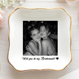 May include: A square white ceramic trinket dish with scalloped edges and a gold-painted rim. The center features a black and white photograph of two people embracing, with the black text "Will you be my Bridesmaid?" and a small heart icon below.