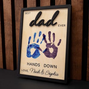 May include: A framed wooden sign with the words "BEST dad EVER" in black 3D lettering. Below are two handprints, one blue and one purple, with the text "HANDS DOWN LOVE, Noah & Sophie".