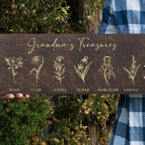 Puede incluir: Un letrero de madera marr&oacute;n oscuro con el texto "Grandma's Treasures" en letras doradas. Seis flores diferentes est&aacute;n grabadas en el letrero, cada una con un nombre debajo: Noah, Clair, Sophia, Elijah, Marceline y Lindsay.