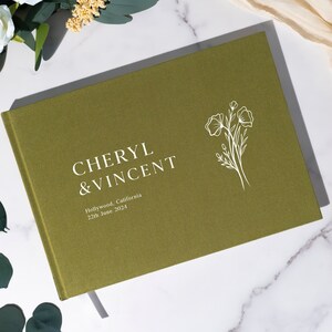 Puede incluir: Libro de visitas de boda verde oliva con los nombres "Cheryl & Vincent" en letras blancas. El libro tambi&eacute;n incluye el lugar y la fecha de la boda. Un dise&ntilde;o floral blanco est&aacute; en el lado derecho.