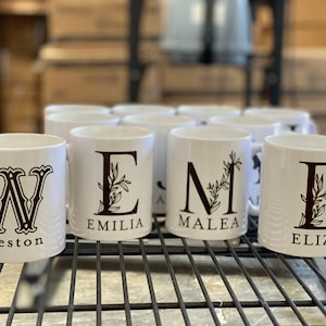 May include: A set of six white ceramic mugs with black lettering. Each mug features a different initial with a floral design and a name below. The names are Weston, Emilia, Malea, Eliza, and two other names that are not visible in the image.