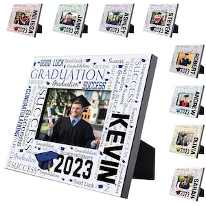 May include: A white picture frame with a black stand. The frame is decorated with the words "Good Luck", "Congratulations", "Graduation", and "Success" in blue and black text. The year "2023" is printed in black on the bottom right corner of the frame. The frame is designed to hold a 4x6 inch photo.