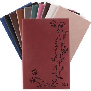 May include: A burgundy leather journal with a floral design and the name "Sophia Harrison" and the year "2023" engraved on the front cover.