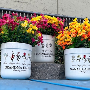 May include: Three white ceramic flower pots filled with colorful flowers. The pots are decorated with floral illustrations and text that reads "Grandma Elsie's Blooming Garden", "Grandma's Garden", and "Nana's Garden we love you".