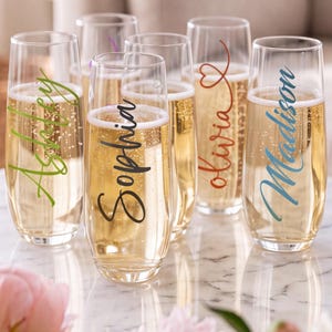 May include: A set of five stemless champagne flutes filled with sparkling liquid. Each glass features a name printed in a unique color and script font: Ashley in green, Sophia in black, Olivia in red with a heart, and Madison in blue.