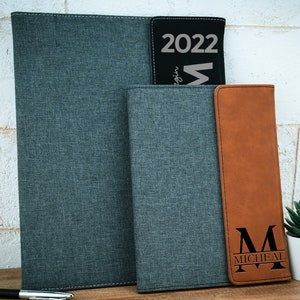 May include: Three different personalized leather-bound journals. The first journal is gray with a textured fabric cover. The second journal is gray with a textured fabric cover. The third journal is brown with a textured leather cover and the letter "M" with the name "MICHEAL" engraved on the front.