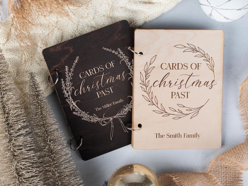 May include: Two wooden photo albums with a ring binding. The albums are decorated with a wreath design and the text "Cards of Christmas Past". One album is dark brown and the other is light brown. The text "The Miller Family" is on the dark brown album and "The Smith Family" is on the light brown album.