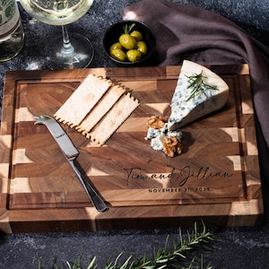 May include: A patterned wooden chopping board with cheese, crackers, olives, and a cheese knife. The board is engraved with the names "Tim and Jillian" and the date "November 21, 2025". Wine glasses and a bottle of wine are also present.
