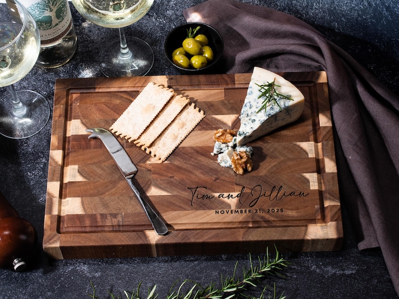 May include: A patterned wooden chopping board with cheese, crackers, olives, and a cheese knife. The board is engraved with the names "Tim and Jillian" and the date "November 21, 2025". Wine glasses and a bottle of wine are also present.