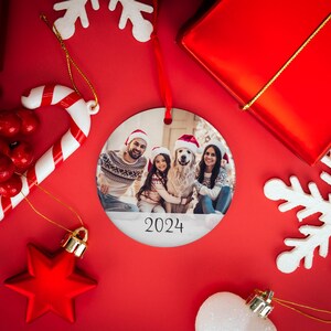 May include: A round white Christmas ornament with a photo of a family with their dog. The family is wearing Santa hats and the dog is wearing a red Santa hat. The ornament has the year 2024 printed on it.