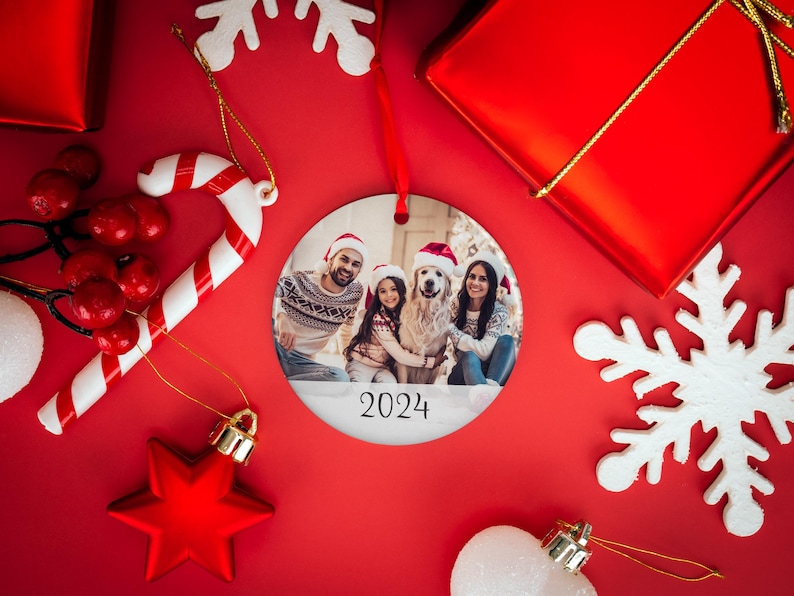 May include: A round white Christmas ornament with a photo of a family with their dog. The family is wearing Santa hats and the dog is wearing a red Santa hat. The ornament has the year 2024 printed on it.