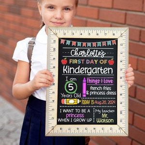 Puede incluir: Un letrero de pizarra con un marco de madera y una regla en el lateral. El letrero dice "Charlotte's First Day of Kindergarten" e incluye detalles como su edad, cosas favoritas y el nombre de su maestro. El letrero est&aacute; decorado con dibujos y texto coloridos.
