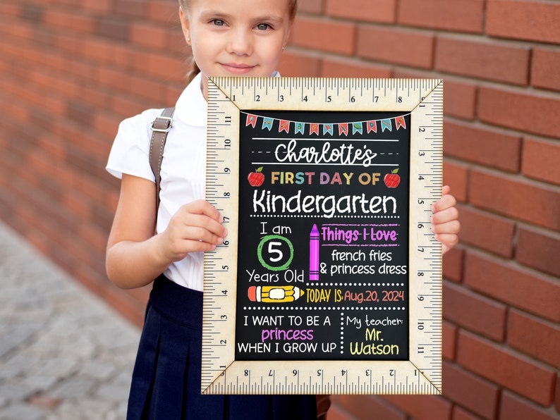 Puede incluir: Un letrero de pizarra con un marco de madera y una regla en el lateral. El letrero dice "Charlotte's First Day of Kindergarten" e incluye detalles como su edad, cosas favoritas y el nombre de su maestro. El letrero est&aacute; decorado con dibujos y texto coloridos.
